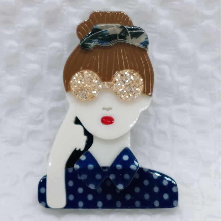 Broche en acrylique femme, chignon brun, serre-tête bleuté, lunettes jaunes pailletées, chemise bleue à pois