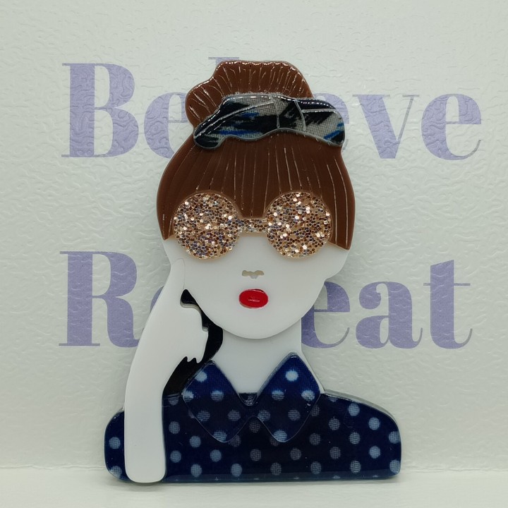 Broche en acrylique femme, chignon brun, serre-tête bleuté, lunettes jaunes pailletées, chemise bleue à pois